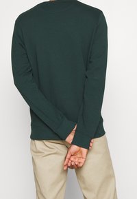 Sudadera de algodón verde oscuro con mangas largas, cuello redondo y puños acanalados. Lleva puestos pantalones beige claros, con las manos entrelazadas detrás.