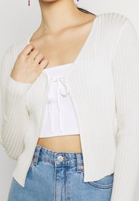Bílý žebrovaný kardigan s výstřihem do V a dlouhými rukávy, s krátkou délkou a otevřeným předním dílem, doplněný o sladěný top.