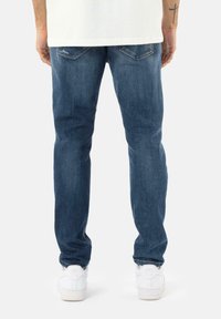Jeans de mezclilla azul oscuro con corte recto, decoloración sutil, bolsillos traseros y detalles desgastados. Combinados con zapatillas blancas.