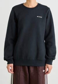 Felpa nera in misto cotone, con girocollo, polsini e fondo a coste. Un piccolo patch con il logo del brand è visibile sul petto.