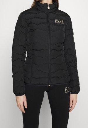 Femme portant une veste matelassée noire EA7 Emporio Armani zippée et des leggings noirs assortis avec le logo EA7 sur la cuisse supérieure.