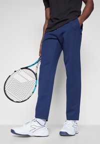 Marineblå treningsbukser med jevn tekstur og sidelommer, kombinert med hvite tennissko og et svart tennisracket.