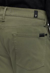 Pantaloni verde oliva con una tasca posteriore, cuciture decorative e un'etichetta nera con il numero "7". Texture liscia, design aderente.