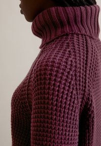 Gebreide pullover in diep paars met een hoge, ribbelige kraag en een textuurpatroon, met een chunky, warme uitstraling.