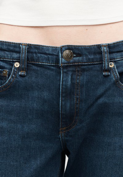 Κοντινό πλάνο μέσης που φοράει μπλε τζιν με μπρούτζινο κουμπί και πορτοκαλί ραφές, σε συνδυασμό με λευκό τοπ που είναι μέσα από το παντελόνι.