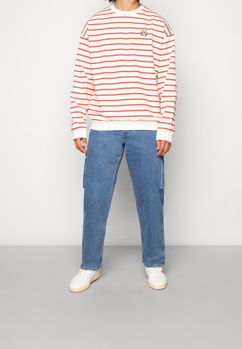 Personne portant un t-shirt à manches longues blanc et rouge à rayures, un jean bleu et des baskets blanches, debout devant un fond uni.