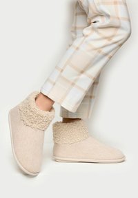 Beige pantofle s texturovaným tkaným svrškem a huňatým lemem. Měkký materiál; nízká výška; hladká, flexibilní podrážka. Spárováno s světlými kostkovanými kalhotami.