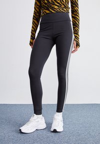 Černé legíny s vysokým pasem, zdobené bílými bočními pruhy a hladkým povrchem. Nosené s bílými sportovními botami na modrém koberci.