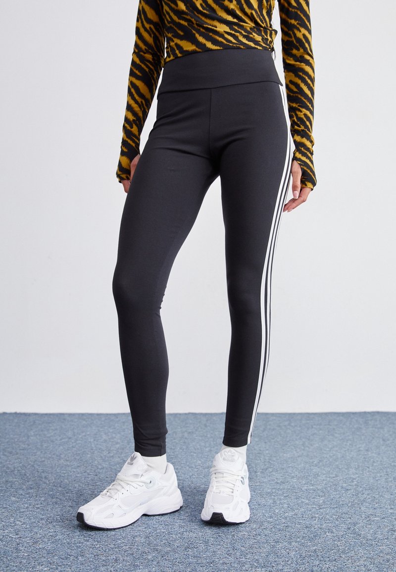 Černé legíny s vysokým pasem, zdobené bílými bočními pruhy a hladkým povrchem. Nosené s bílými sportovními botami na modrém koberci.