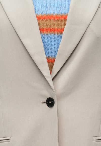 Ανοιχτό μπεζ σακάκι με μαύρο κουμπί, σε συνδυασμό με μπλε πλεκτό πουλόβερ με οριζόντιες ρίγες σε πορτοκαλί, καφέ και μπεζ.