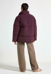 Giubbotto imbottito viola con colletto alto, caratterizzato da un design trapuntato. Abbinato a pantaloni larghi di un marrone chiaro e sneakers viola.