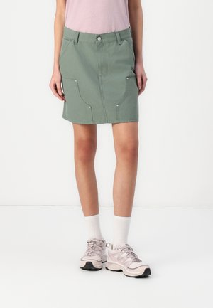 Femme portant une jupe verte au genou avec des poches avant, des chaussettes blanches et des chaussures de sport rose clair, debout sur un fond blanc.