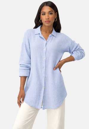 Frau mit langen dunklen Haaren, die goldene Creolenohrringe trägt, ein hellblaues strukturiertes Hemd mit geknöpften Ärmeln und weißen Hosen, posiert mit der Hand auf der Hüfte.