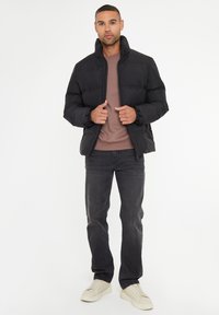 Zwarte pufferjack met een hoge kraag, voorzien van een ritssluiting en elastische manchetten. Draagt over een bruine lange mouwen shirt en grijze jeans.