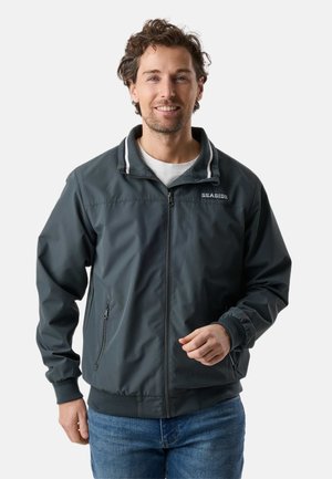 Mężczyzna z kręconymi włosami, ubrany w ciemnoszarą kurtkę na zamek z napisem "SEASIDE" i niebieskie dżinsy, uśmiechający się na jednolitym tle.