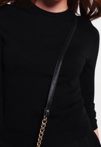 Maglione nero a maglia con scollatura tonda, maniche aderenti e una texture liscia. È visibile una tracolla nera con dettagli in catena dorata.