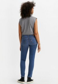 Jeans ajustados de mezclilla de cintura alta, mostrados desde la parte de atrás, combinados con una blusa sin mangas gris y zapatillas negras.