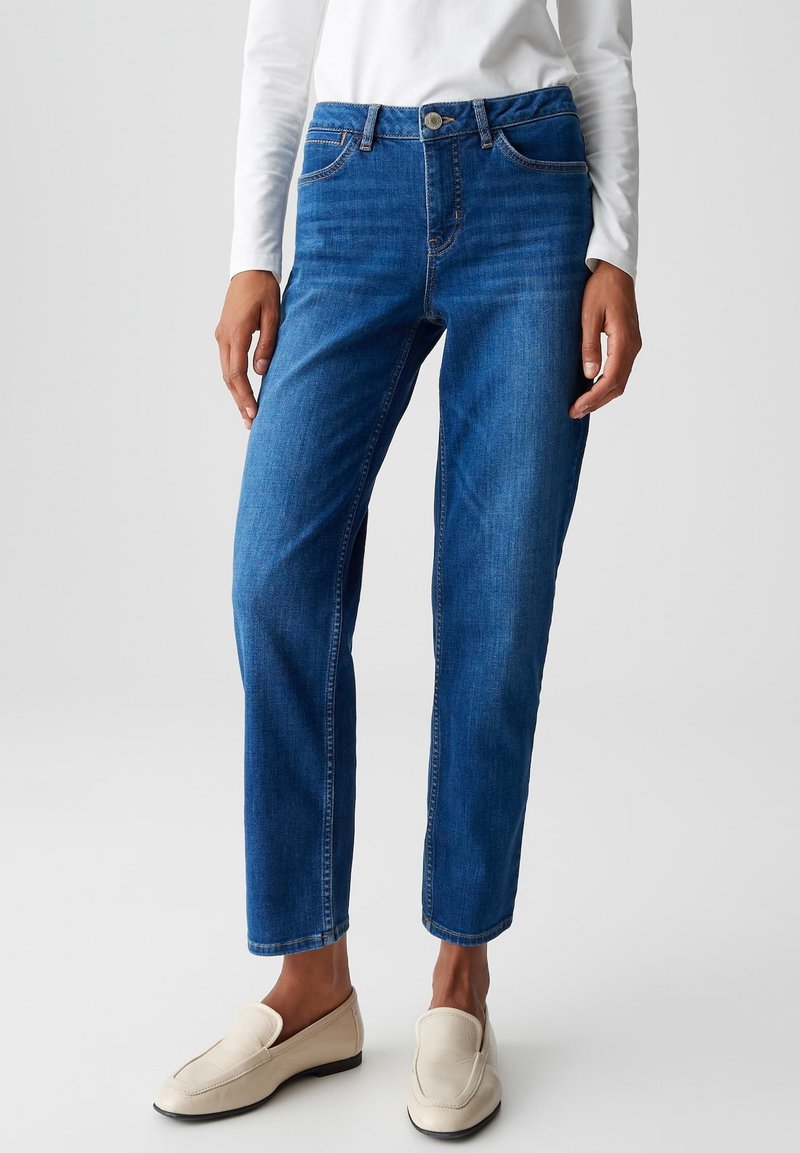 Personne portant un jean droit bleu, un haut à manches longues blanc et des mocassins beiges, se tenant devant un fond clair uniformément lumineux.