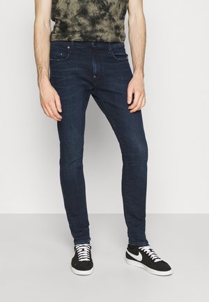 Mężczyzna w dopasowanych ciemnoniebieskich dżinsach, czarno-białych trampkach i zielono-czarnej koszulce tie-dye, stojący na tle jednolitego jasnego tła.