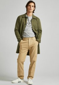 Abrigo verde oliva con botones, camiseta gris con logo, pantalones beige y zapatillas blancas. Tejido suave, corte casual y diseño sencillo.