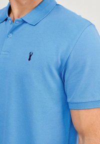 Jasnoniebieska koszulka polo z teksturowanej tkaniny, z wykładanym kołnierzykiem i małym czarnym emblematem homara na piersi.