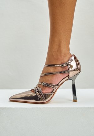 Escarpin à talon haut avec une finition métallisée rose doré, bout pointu, plusieurs fines lanières, boucles décoratives et un talon aiguille noir brillant.