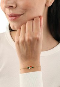 Kvinna som bär ett delikat guldarmband med gröna, röda och vita marquise-formade stenar, vilande hand nära hennes ansikte.