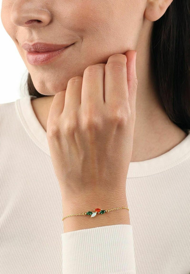 Kvinna som bär ett delikat guldarmband med gröna, röda och vita marquise-formade stenar, vilande hand nära hennes ansikte.