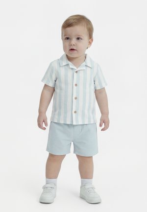 Dijete stoji, nosi plavu i bijelu prugastu majicu kratkih rukava, svijetloplave kratke hlače, bijele čarape i bijele tenisice.