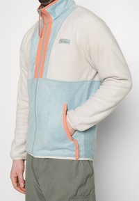 Giacca in fleece leggera con un design a blocchi di colore blu e crema, colletto alto, chiusura con zip e dettagli arancioni su zip e tasche.