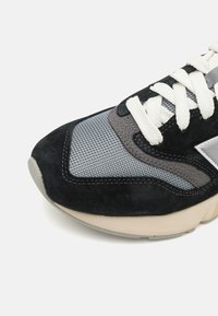 Adidași negri și gri, cu un guler din plasă, accente din piele întoarsă, șireturi albe și o talpă texturată. Designul se concentrează pe confort și susținere.