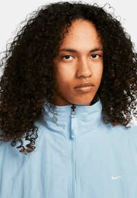 Giacca zip-up azzurra chiara con colletto alto, realizzata in tessuto leggero con una sottile trama e un logo Nike bianco sul lato sinistro del petto.