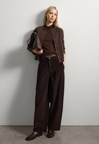Cardigán de punto marrón sobre una blusa marrón, combinado con pantalones acampanados de pana. Bolso de mano texturizado marrón en la mano, combinando con mocasines marrones.