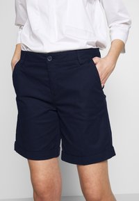 Mörkblå bomullsshorts med en jämn textur, vikta fållar, framfickor och en knäppning vid midjan. Bärs tillsammans med en vit blus.