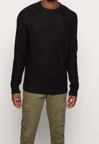 Homme portant un pull noir à manches longues côtelé et un pantalon cargo vert olive avec poches à boutons, debout devant un fond clair uni.