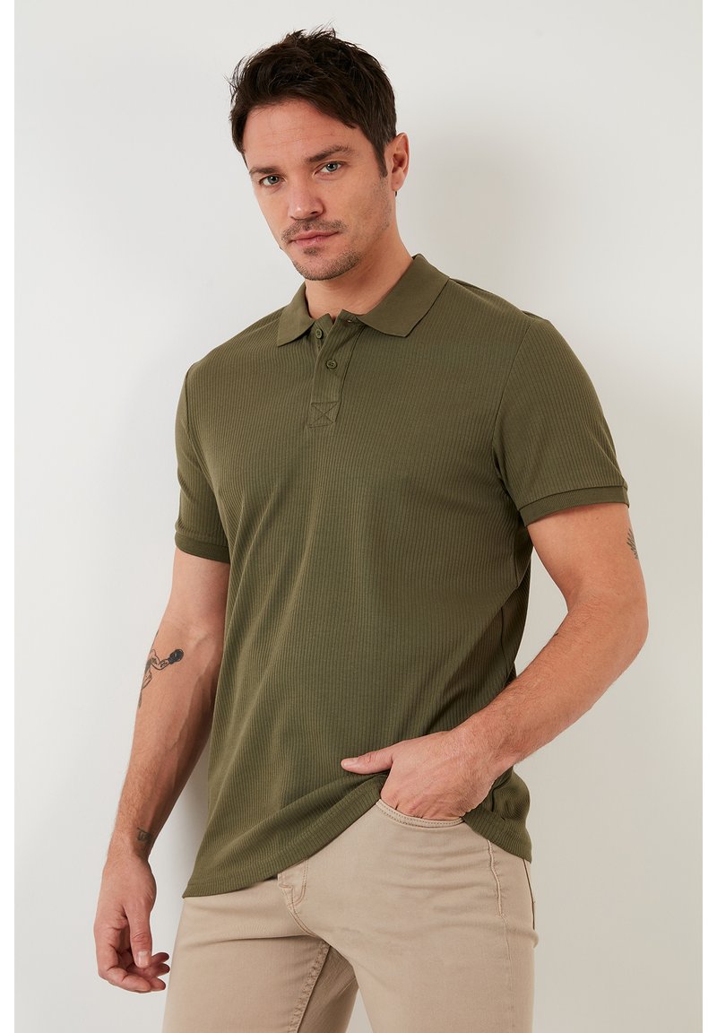 Camisa polo de canalé en verde oliva con cuello, dos botones y mangas cortas, combinada con pantalones de color beige claro. Presentada sobre un fondo liso.