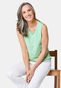 Mouwloze groene blouse van zacht materiaal, ronde halslijn, gecombineerd met witte broek. Glad textuur met een ontspannen pasvorm, model zittend op een houten stoel.