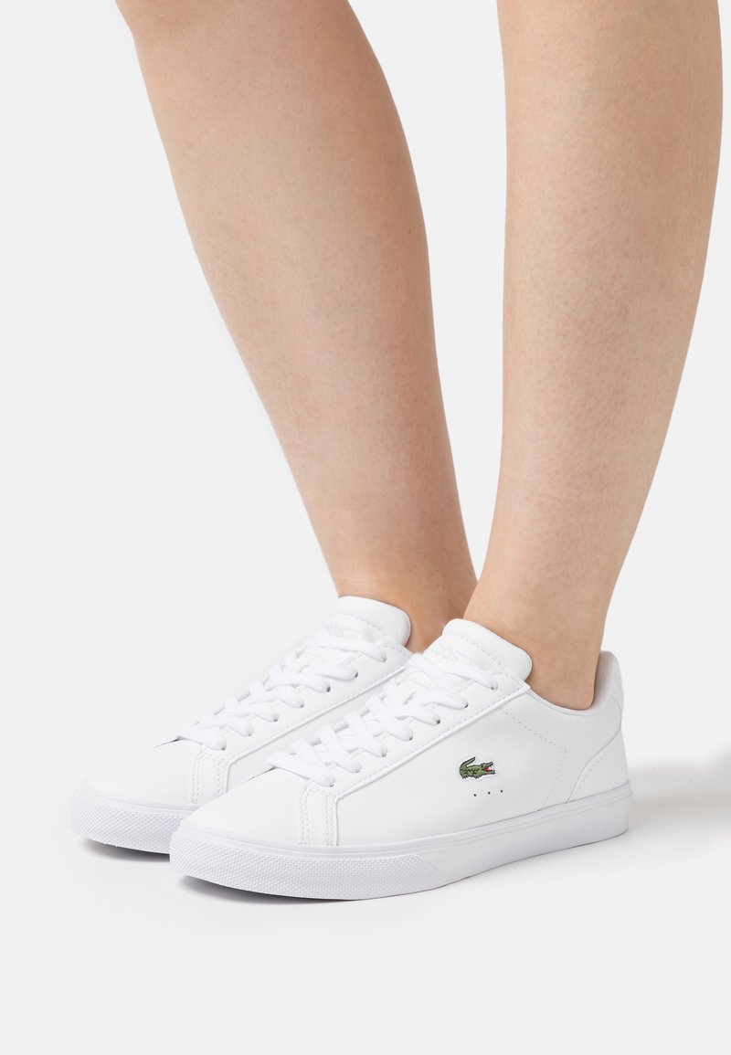White sneakers made of smooth leather with round toe, flat rubber sole, lace-up front, and a small green crocodile logo on the side.