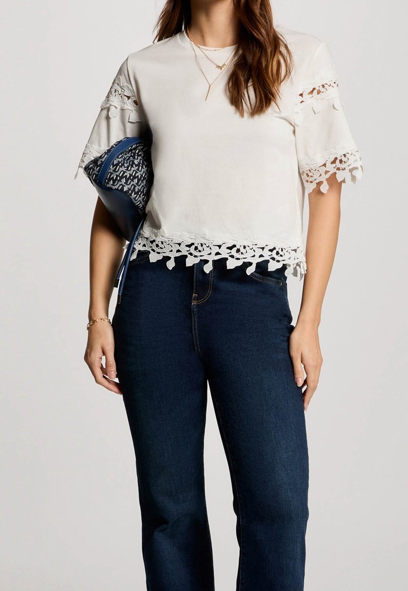 Femme portant un chemisier blanc à manches courtes avec bordure en dentelle et un jean bleu, tenant une pochette bleue à motifs sous le bras.