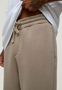 Beige sweatpants met een drawstring tailleband, zijzakken en een zachte textuur, gecombineerd met een witte t-shirt die gedeeltelijk omhoog is getrokken.
