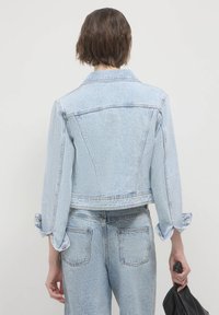 Giacca di jeans azzurro chiaro con design corto, maniche lunghe e polsini abbottonati. Presenta un colletto classico e dettagli cuciti.