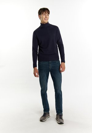 Tengerészt navy turtleneck pulóver kötött anyagból, kék farmernadrággal párosítva. A modellen szürke sportcipő van, amelynek díszítése és textúrája is van.