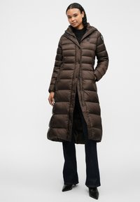 Brun pufferjakke med hætte, med en quiltet design, trykknaplukninger og en lang længde. Båret over mørke jeans og hælede sko.