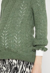 Pulôver verde de malha com um intrincado padrão de folhas, tecido texturizado e detalhes de punhos franzidos. Usado com uma saia de estampa de zebra em preto e branco.