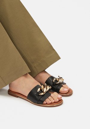 Sandali scivolati in pelle nera con una larga fascia, impreziositi da una catena dorata, suola piatta e sottopiede color marrone chiaro. Indossati con pantaloni larghi color oliva.