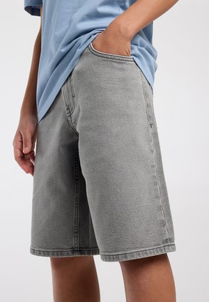 Persona con camisa azul clara y pantalones cortos de mezclilla grises hasta la rodilla, con una mano en el bolsillo sobre un fondo liso.