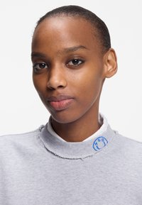 Κοντινό πλάνο μιας γυναίκας με λεία επιδερμίδα, που φοράει ανοιχτό γκρι φουτερ με λευκό γιακά και ένα μικρό μπλε κεντημένο χαμογελαστό κοντά στον λαιμό.