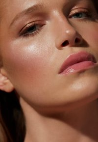 Close-up of a face with smooth, luminous skin. Features include lightly flushed cheeks, full pink lips, and subtle eye makeup.