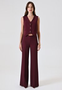 Gilet bordeaux su misura con scollo a V e tre bottoni circolari, abbinato a pantaloni a vita alta e gamba larga con righe verticali.