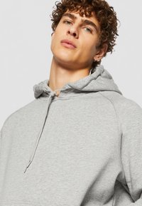 Sudadera gris con cordón, hecha de tela suave. Cuenta con un bolsillo tipo canguro y mangas raglán, mostrando un diseño casual.